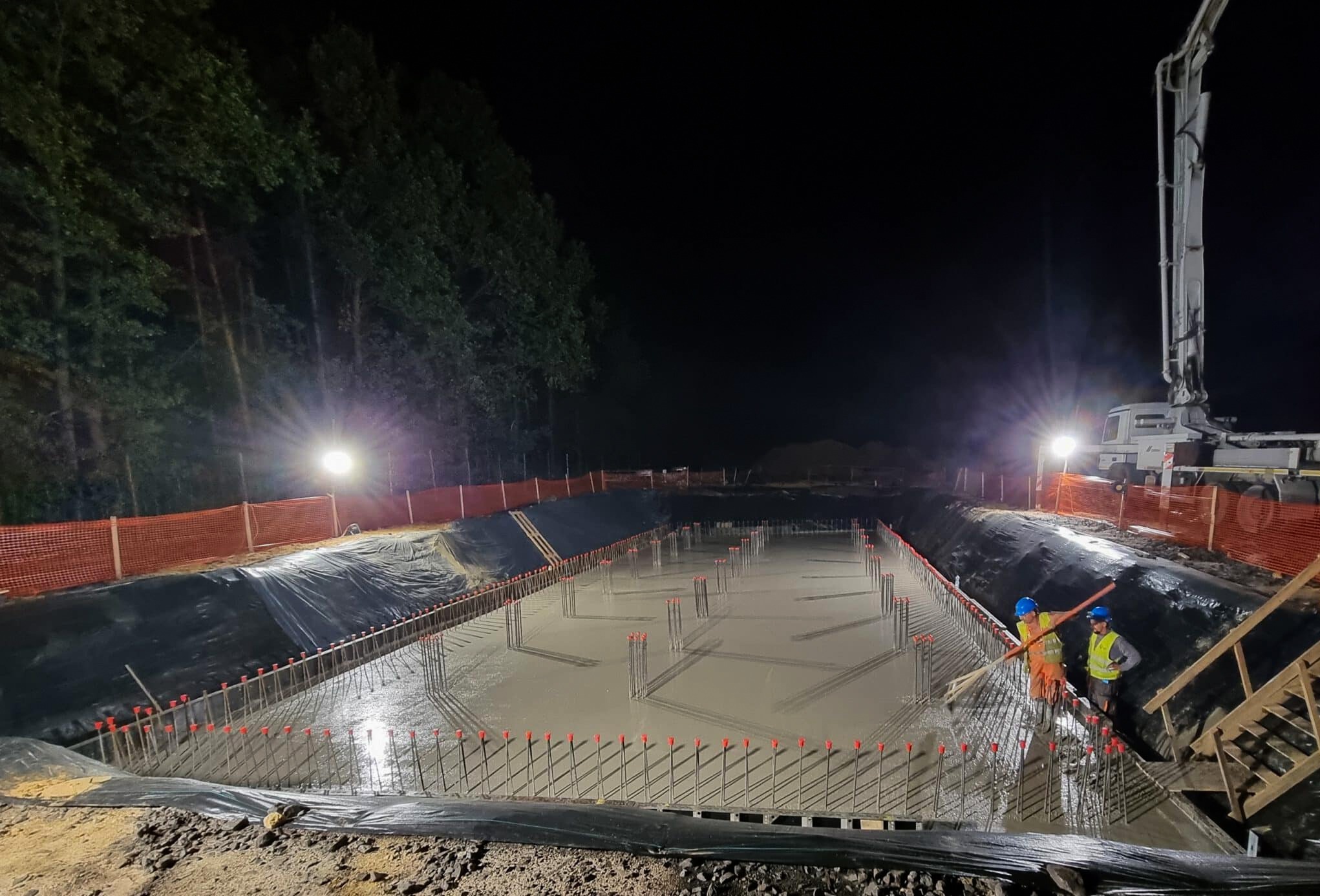 Nocne betonowanie fundamentów zbrojonych na budowie. Widoczni pracownicy w odblaskowych kamizelkach i kaskach, pompa do betonu, oświetlenie budowlane.