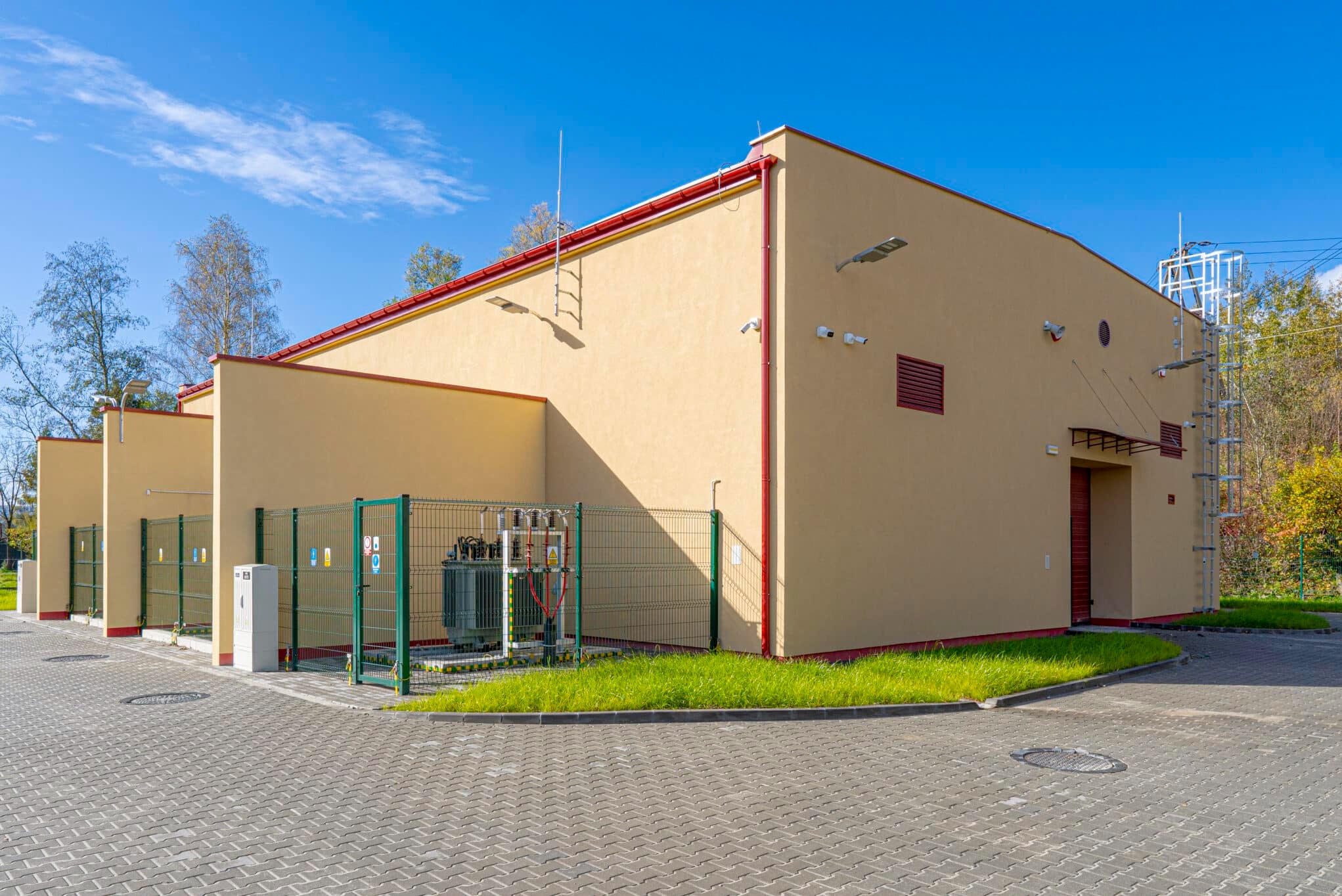 Zewnętrzny widok stacji transformatorowej z zielonym ogrodzeniem i szarym brukiem. Żółty budynek z czerwonymi elementami, oświetlenie zewnętrzne i drabina na dachu.