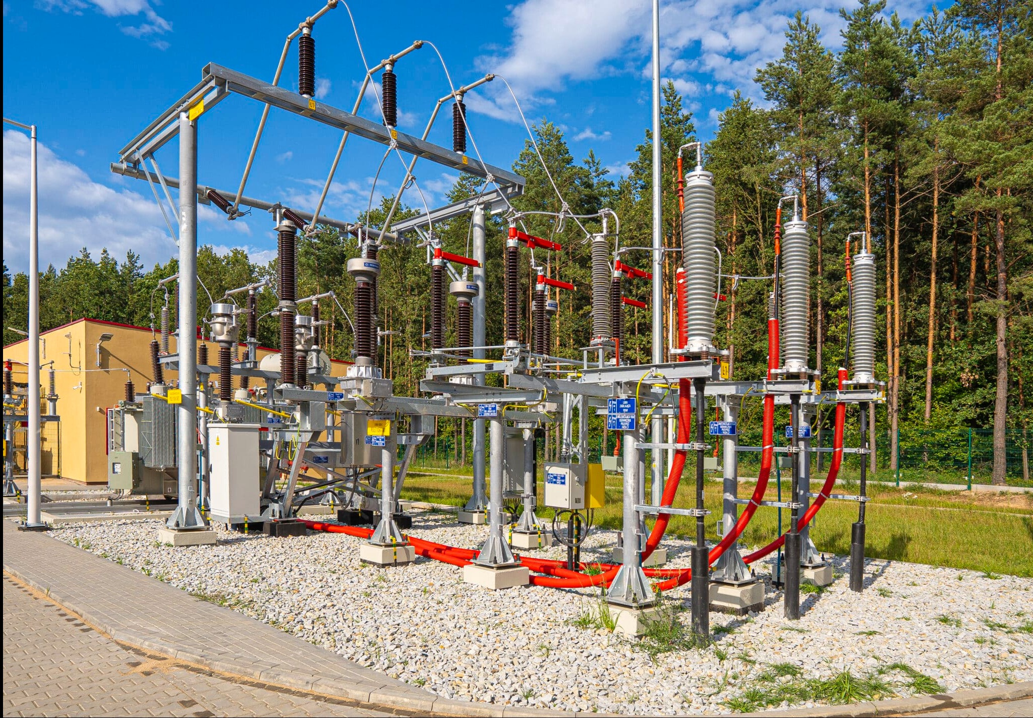 Rozległa stacja transformatorowa z licznymi elementami elektrycznymi na tle lasu i błękitnego nieba. Widoczne transformatory, izolatory i kable.