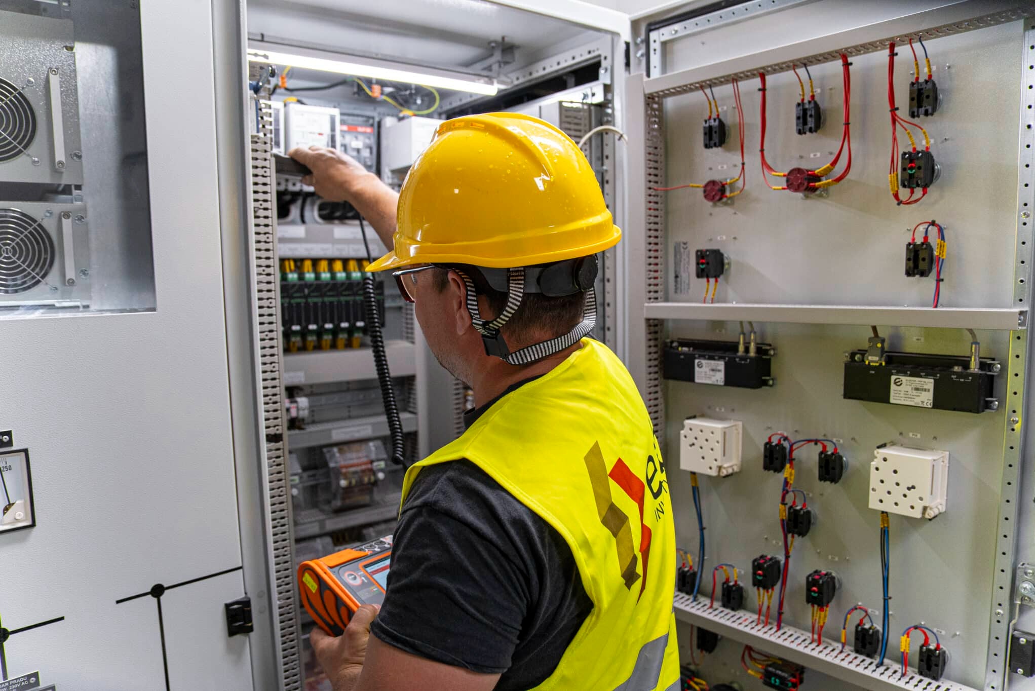 Elektryk w kasku i kamizelce mierzy parametry w szafie sterowniczej. Widoczne przewody, zabezpieczenia i aparatura kontrolno-pomiarowa w rozdzielni elektrycznej.