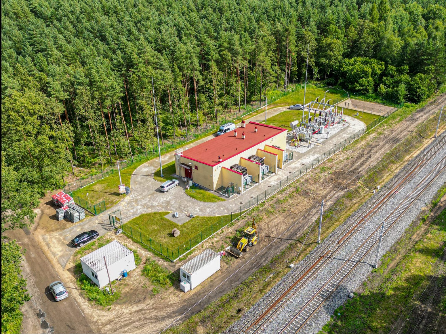 Widok z lotu ptaka na stację transformatorową z czerwonym dachem, otoczoną zielenią i lasem, z linią kolejową w tle. Na terenie stacji zaparkowane samochody i sprzęt budowlany.