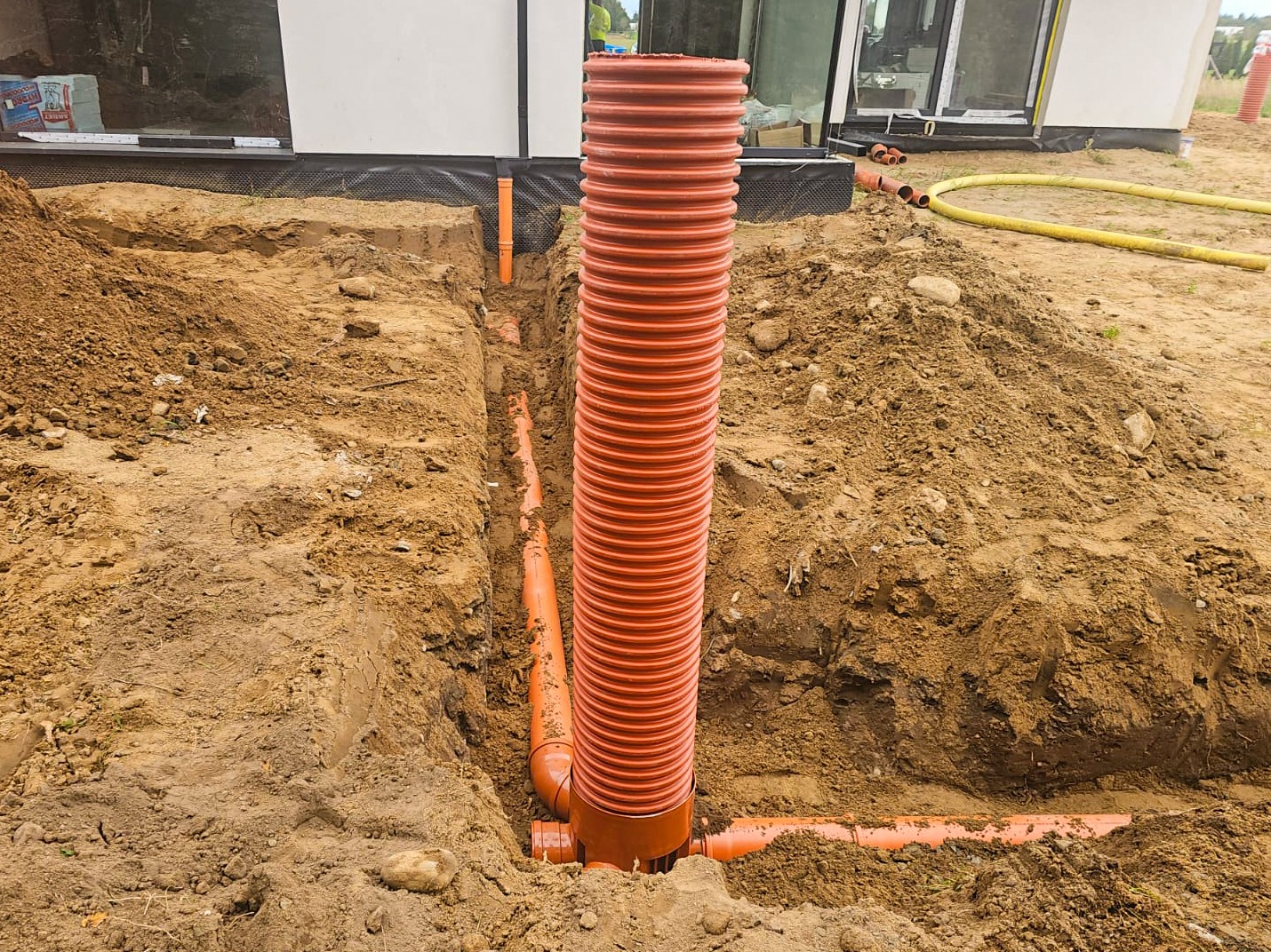 Pomarańczowe rury kanalizacyjne ułożone w wykopie ziemnym przy budynku. Widoczne złącza i pionowa rura karbowana. Prace ziemne w toku, teren budowy.