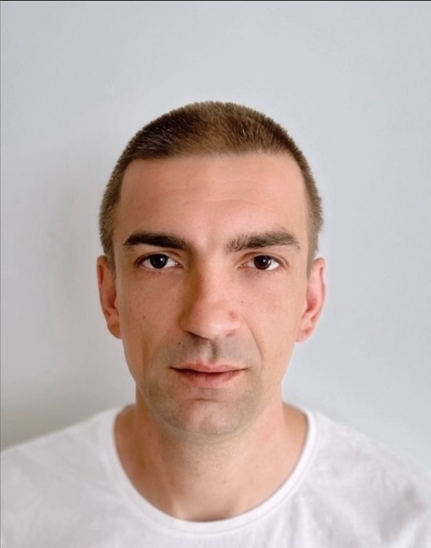 Portret mężczyzny o krótkich włosach, w białej koszulce, na jasnym tle. Neutralny wyraz twarzy, skierowany wprost do kamery. Dobre oświetlenie.