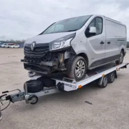 Uszkodzony srebrny Renault Trafic na białej lawecie, holowany przez czarny samochód na parkingu. Widoczne uszkodzenia przodu pojazdu.