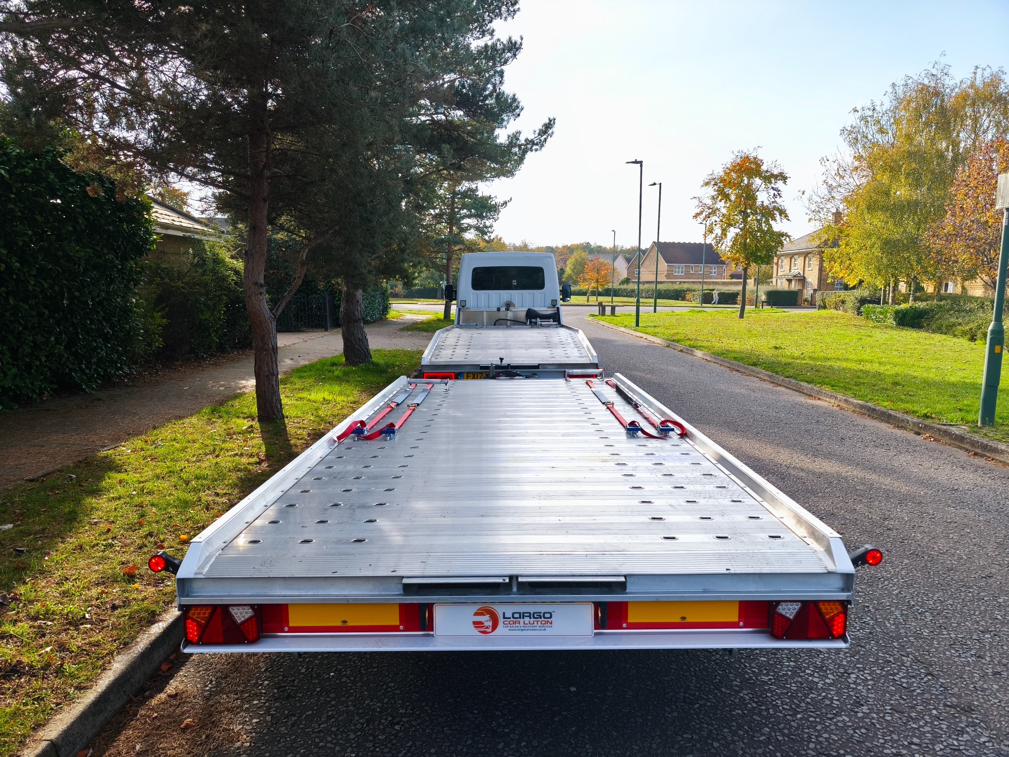 Widok z tyłu lawety samochodowej z białym ciągnikiem, zaparkowanej na asfaltowej drodze w słoneczny dzień. Widoczne mocowania i oświetlenie lawety.