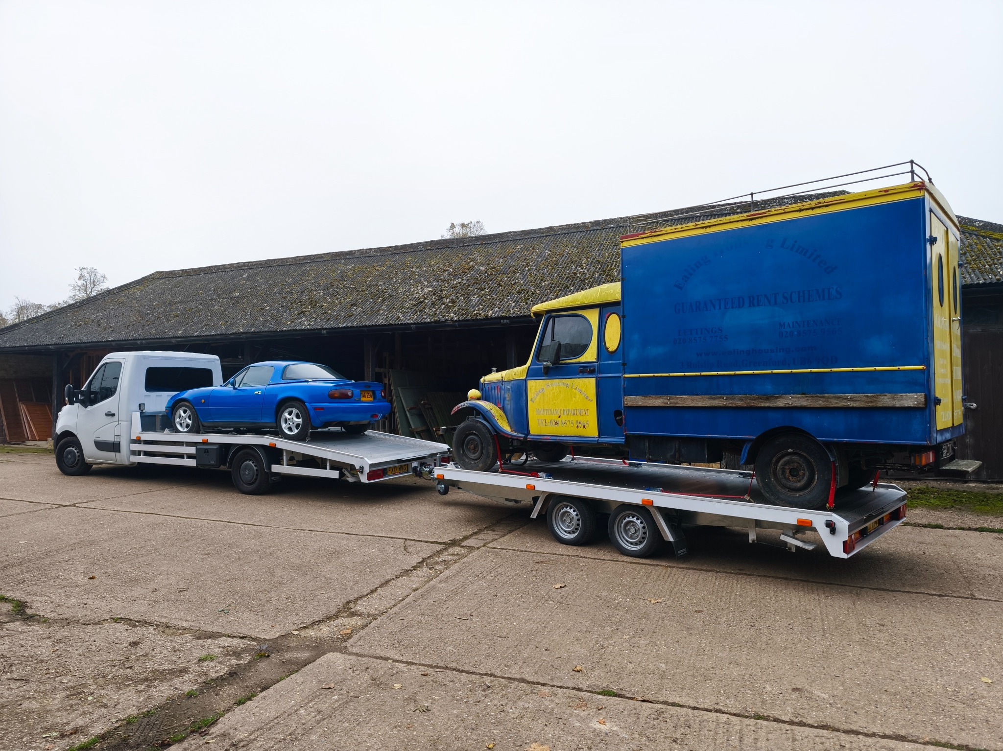 Dwie lawety: biała z niebieską Miatą i niebiesko-żółta z napisem „Ealing Housing Limited”, stoją na betonowym placu przed budynkiem z szarym dachem.