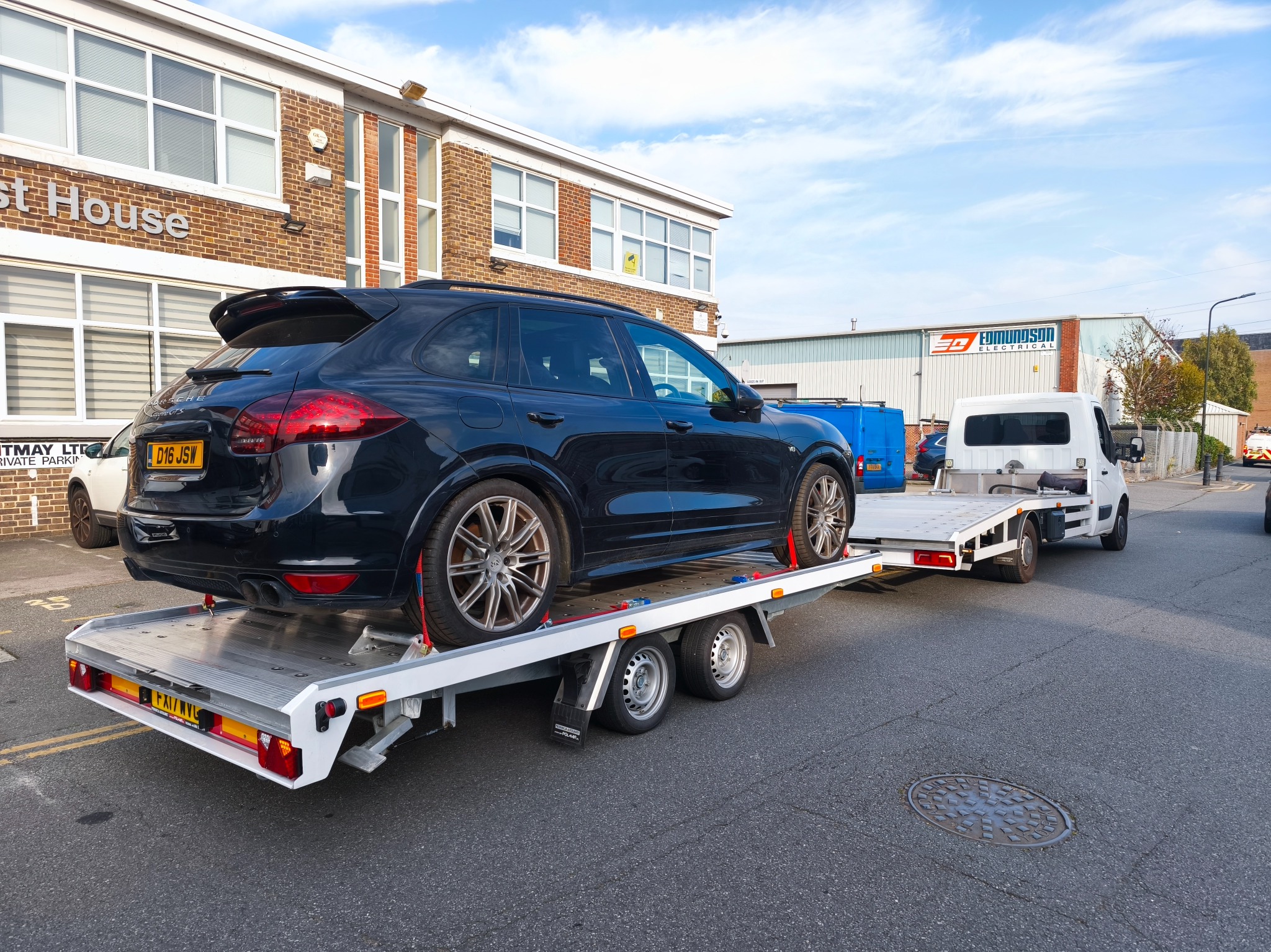 Czarny SUV marki Porsche na niskopodwoziowej lawecie, zabezpieczony pasami transportowymi. W tle budynek i biały samochód dostawczy.