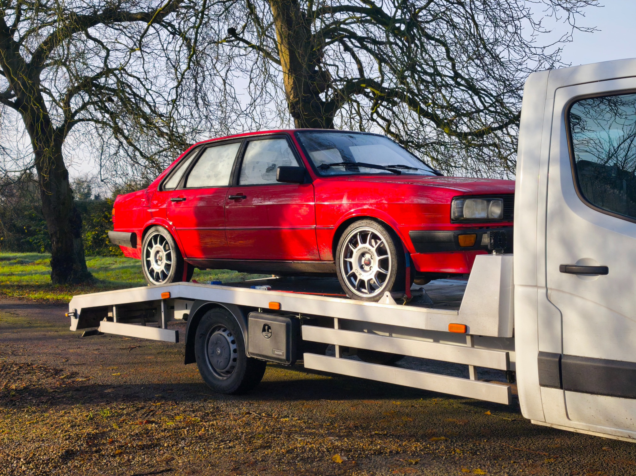 Czerwone Audi 80 na białej lawecie, widok z boku, w tle drzewa bez liści, jesienna sceneria. Transport pojazdu na lawecie.