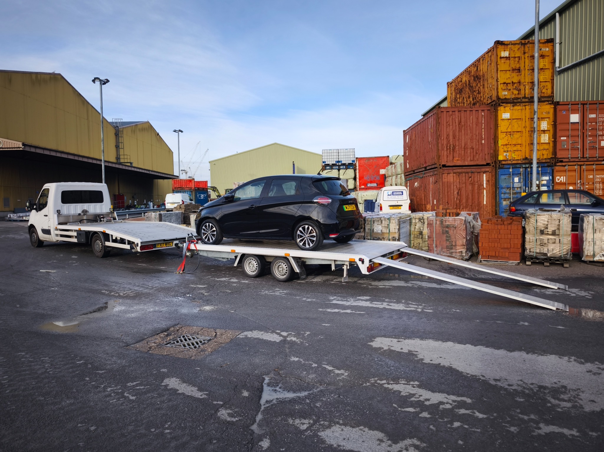 Czarny samochód osobowy na białej lawecie, z rozłożonymi najazdami, na tle magazynów i kontenerów w porcie. Widoczny biały bus ciągnący lawetę.