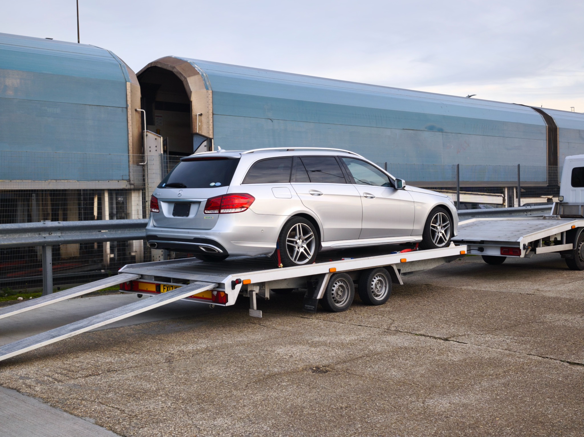 Srebrne kombi Mercedes-Benz na białej lawecie z rozłożonymi najazdami, na tle hali magazynowej z przesuwnymi wrotami.