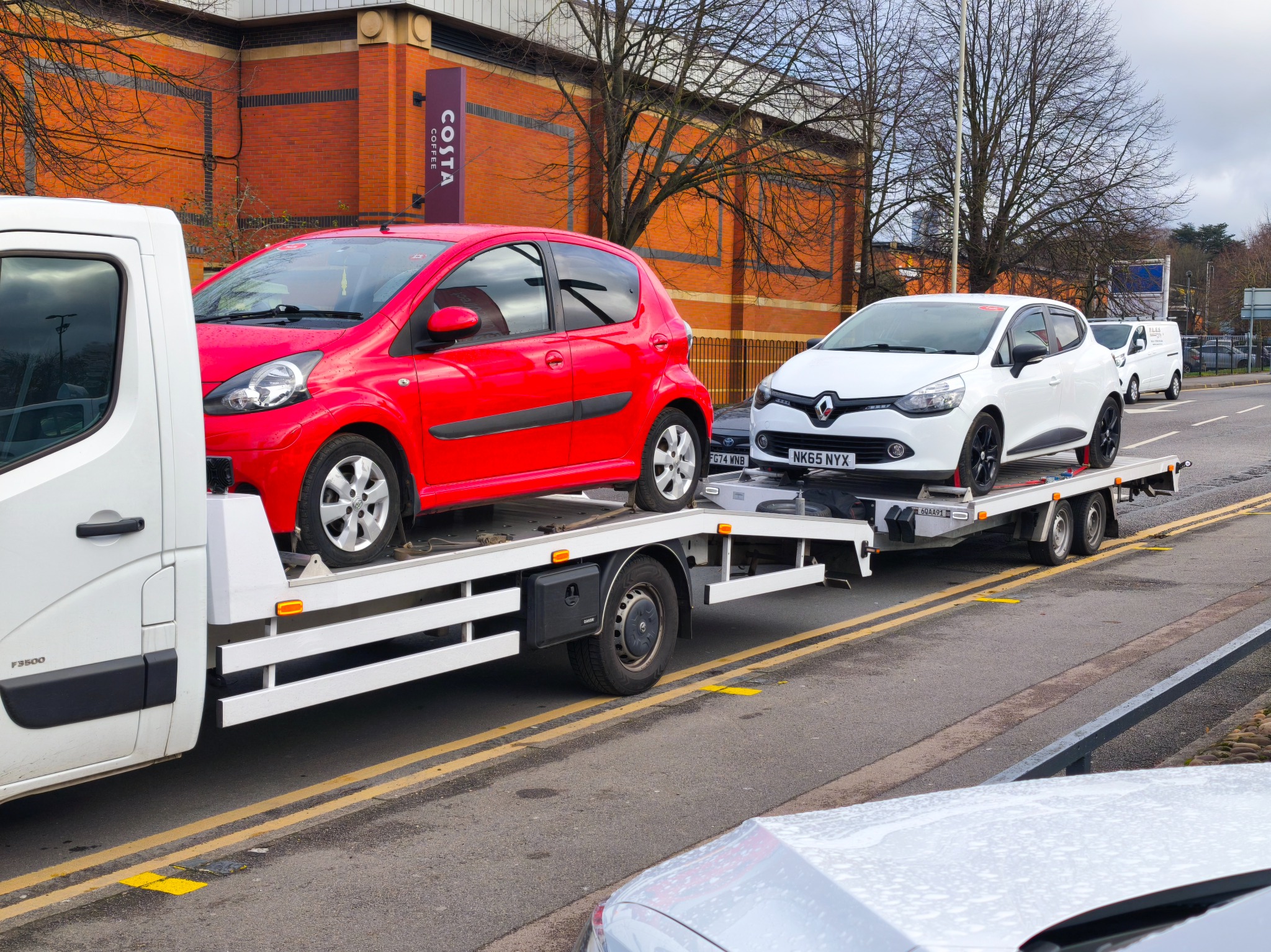 Laweta z czerwonym i białym samochodem na platformie, zaparkowana na ulicy z budynkiem Costa Coffee w tle.