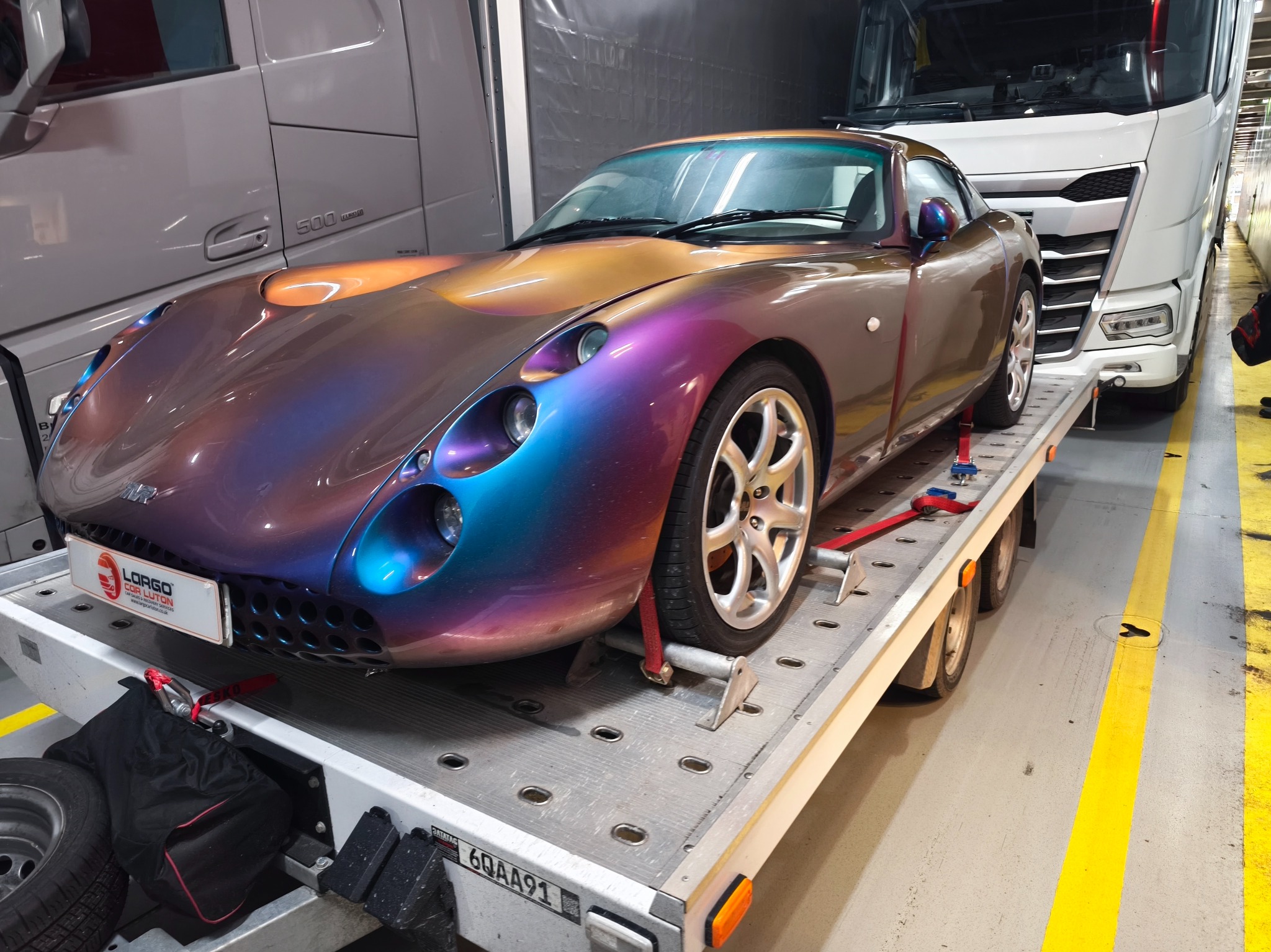 Wielobarwny sportowy samochód na lawecie wewnątrz naczepy transportowej. Auto zabezpieczone pasami transportowymi. Widoczna tablica rejestracyjna lawety.