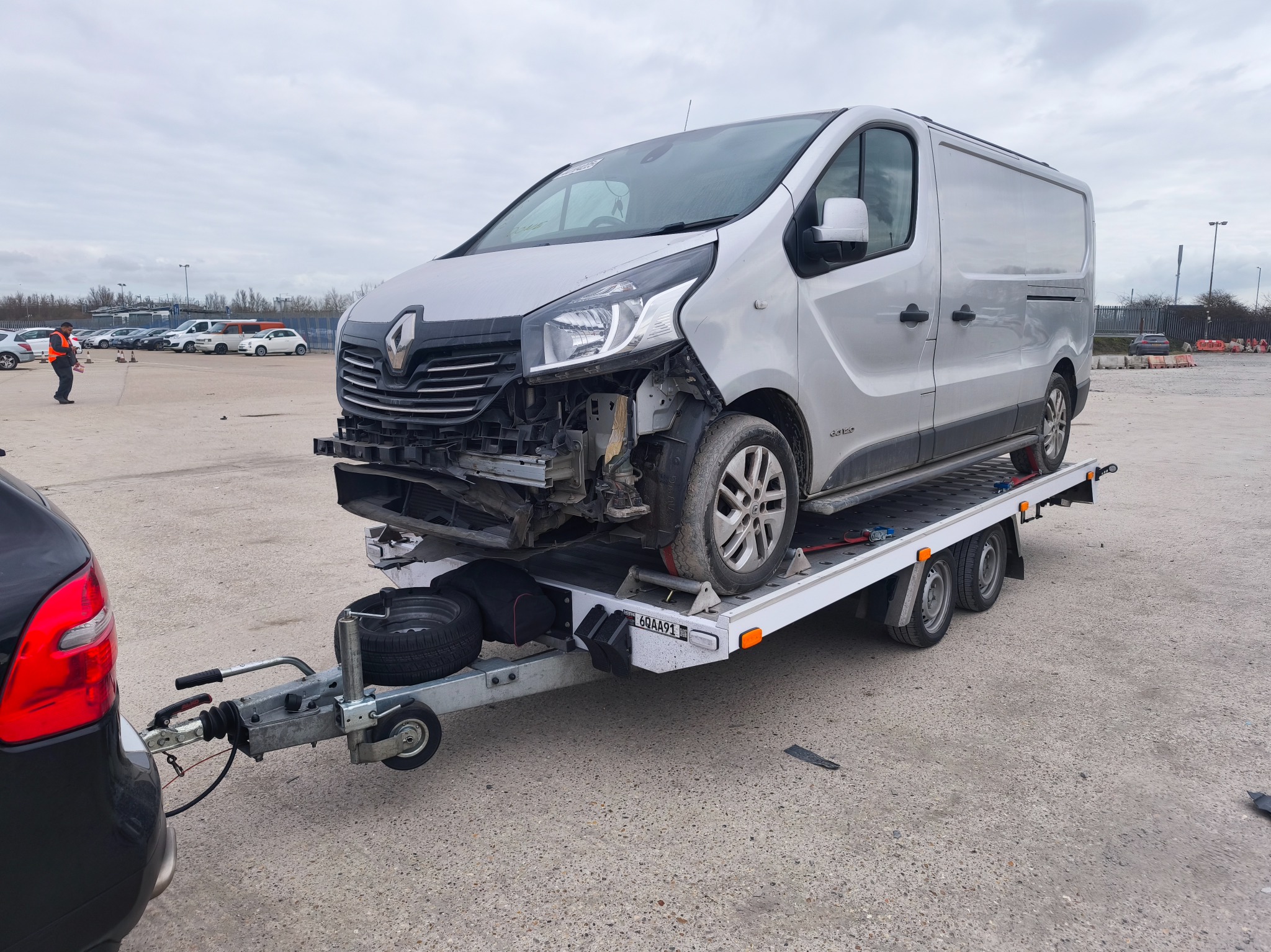 Uszkodzony srebrny Renault Trafic na białej lawecie, holowany przez czarny samochód na parkingu. Widoczne uszkodzenia przodu pojazdu.
