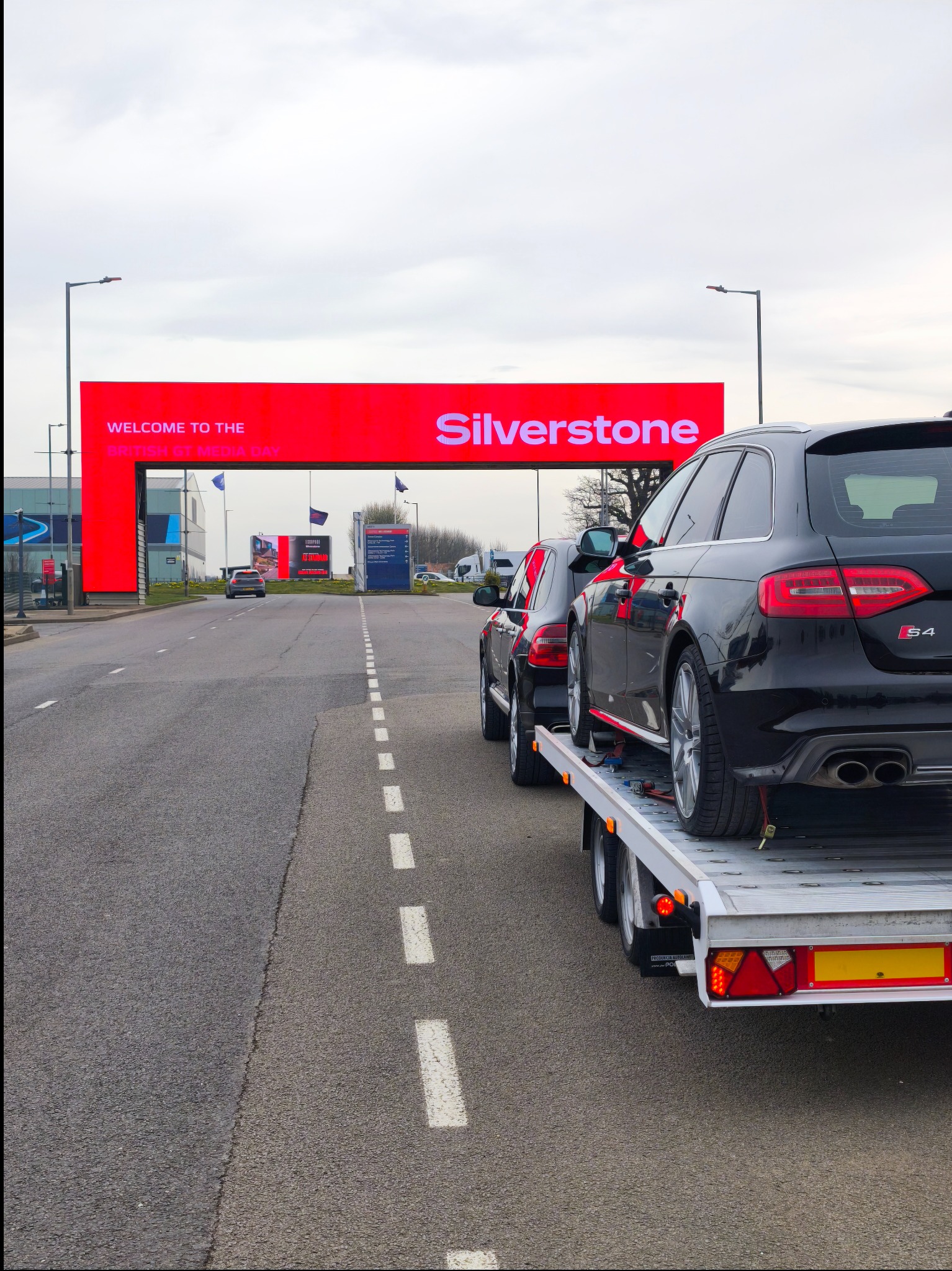Czarny samochód Audi S4 na lawecie przed wjazdem na tor Silverstone. Dwa auta transportowane, widok z tyłu lawety. Tablica powitalna nad wjazdem.