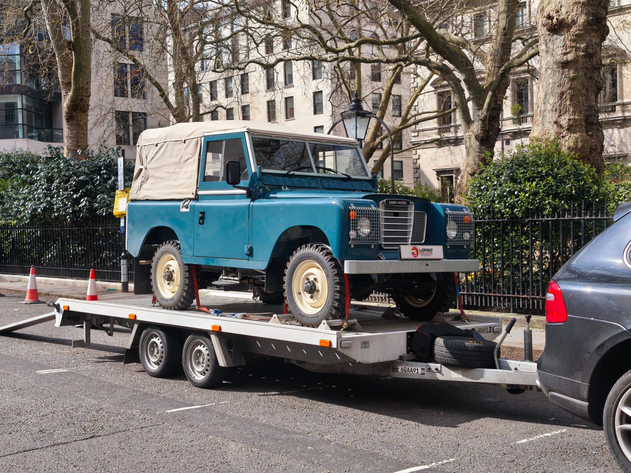 Turkusowy Land Rover na lawecie z podniesioną rampą, zabezpieczony pasami transportowymi na tle miejskiej zabudowy i drzew.