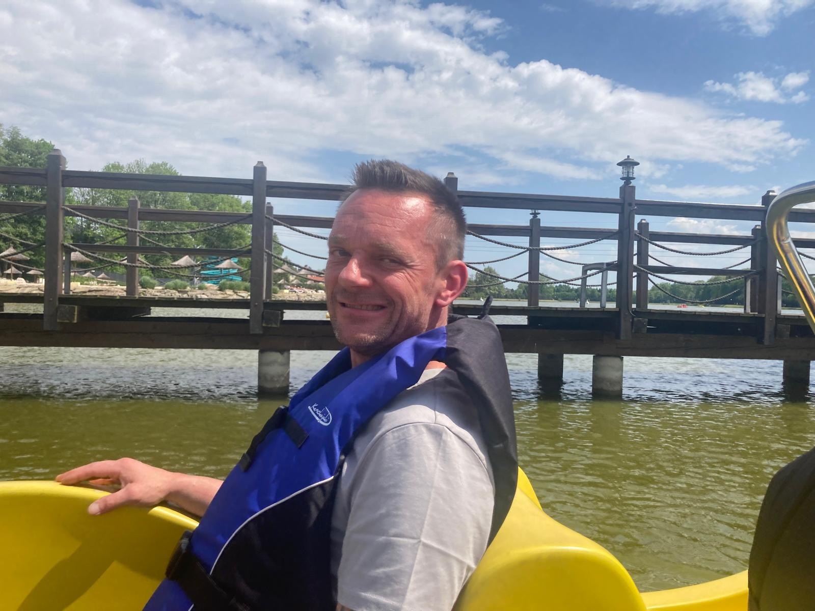 Uśmiechnięty mężczyzna w kamizelce ratunkowej siedzi na żółtym rowerze wodnym na jeziorze, z drewnianym pomostem i błękitnym niebem w tle.