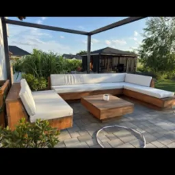 Drewniany narożnik z białymi poduchami na tarasie z kostki, stolik kawowy i pergola w tle. Hula-hop na ziemi dodaje swobodnej atmosfery.