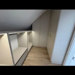 Jasna garderoba na poddaszu ze schowkami i szafami z frontami bez uchwytów. Oświetlenie LED podkreśla nowoczesny charakter wnętrza.