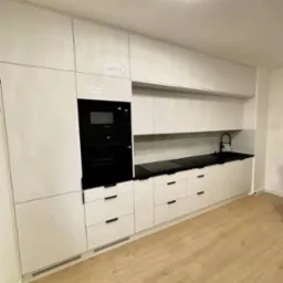 Biała zabudowa kuchenna z czarnym blatem i sprzętem AGD, wykończona drewnianą podłogą i nowoczesnym oświetleniem sufitowym.