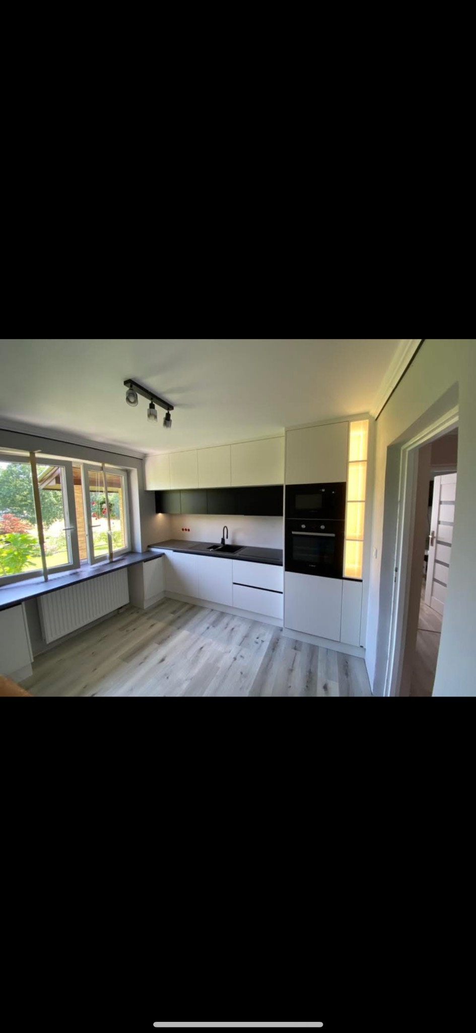 Nowoczesna kuchnia z białymi szafkami, czarnymi blatami i drewnianą podłogą. Duże okno z widokiem na zieleń. Oświetlenie punktowe i listwa LED. Minimalistyczny design.