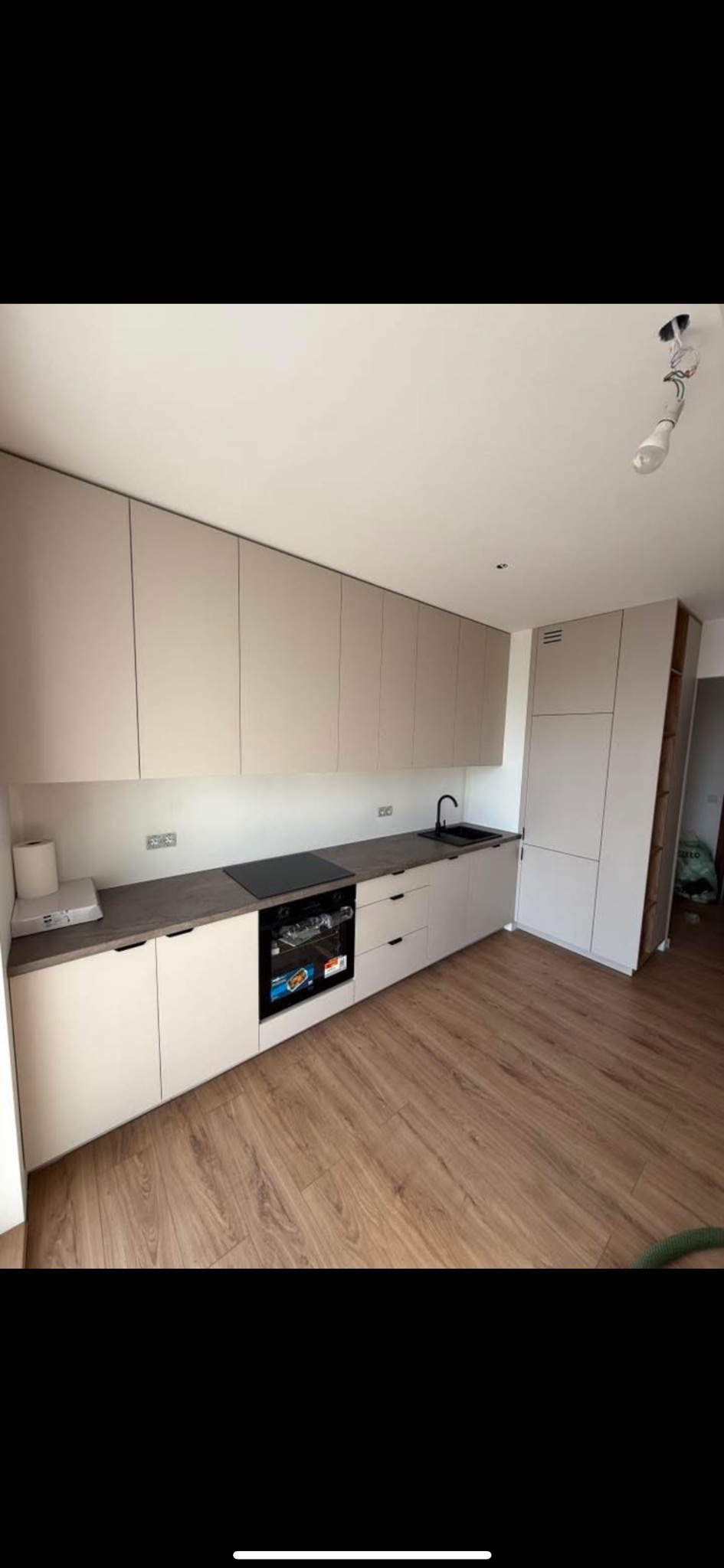 Nowoczesna kuchnia z beżowymi szafkami, ciemnym blatem i drewnianą podłogą. Widoczny piekarnik i czarny zlew. Minimalistyczny design oświetlony naturalnym światłem.