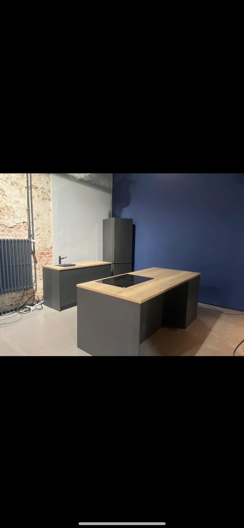 Nowoczesna kuchnia z szarymi szafkami i drewnianymi blatami, z płytą indukcyjną, na tle ceglanej ściany i niebieskiej ściany. Minimalistyczny design, surowe wykończenie.