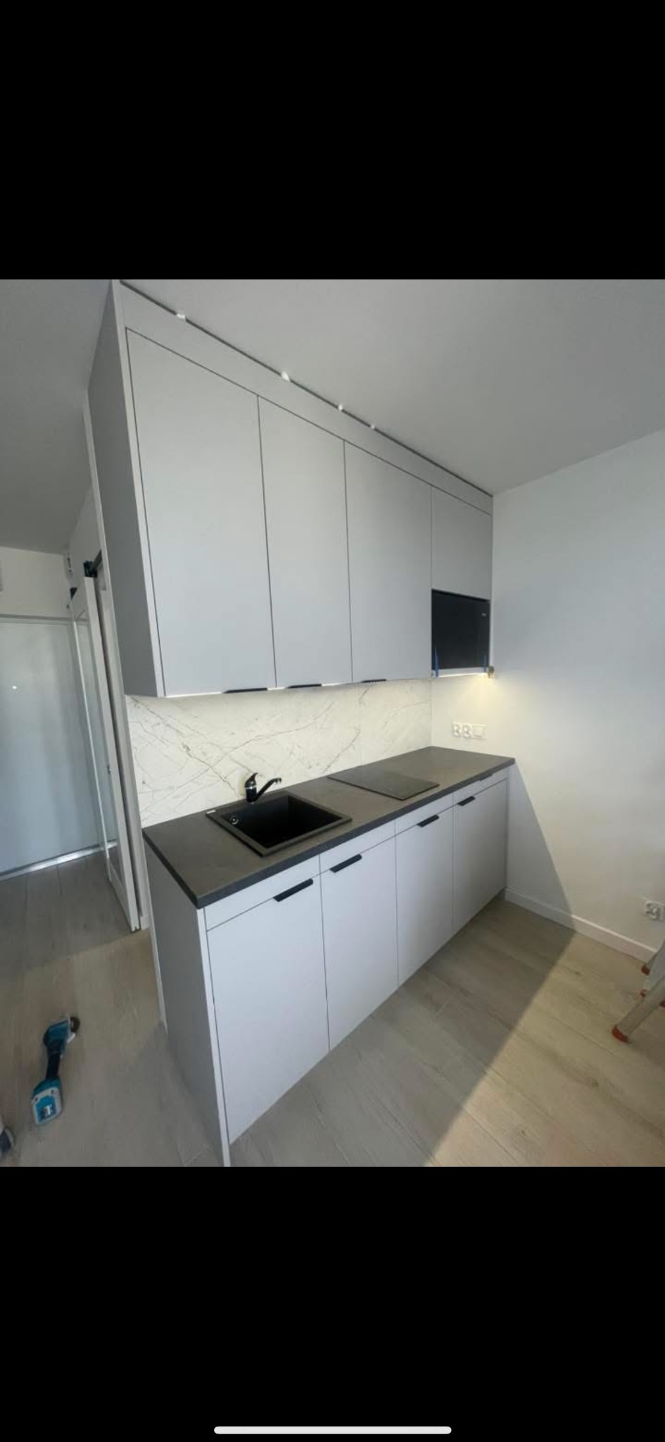 Nowoczesna kuchnia z szarymi szafkami, czarnym blatem i zlewem. Marmurowy backsplash i subtelne oświetlenie podkreślają minimalistyczny design. W tle widoczny sprzęt.