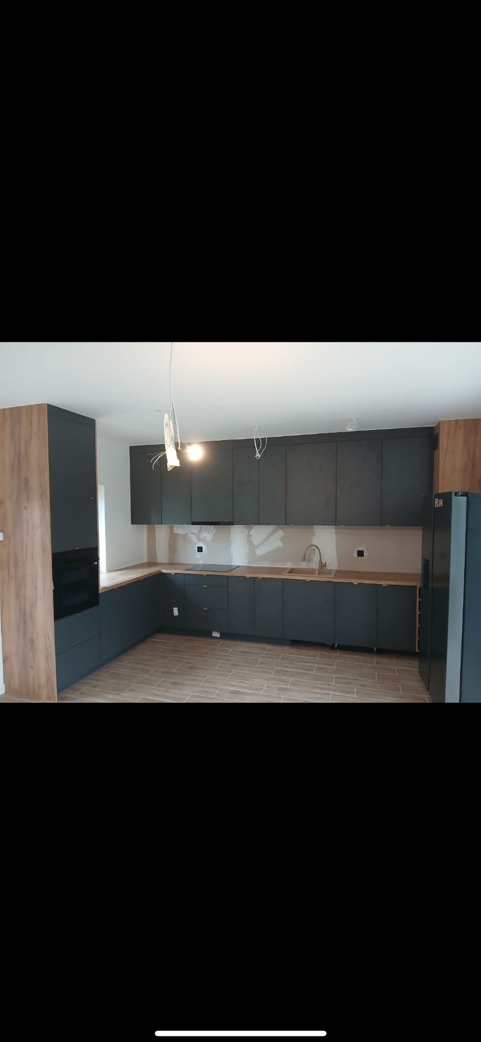 Nowoczesna kuchnia z ciemnymi szafkami i drewnianym blatem. Widoczne niedokończone wykończenia ścian i podłączenia elektryczne.