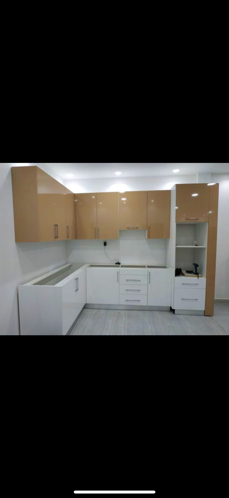 Montaż kuchni na wymiar: białe dolne szafki z szufladami i beżowe górne szafki z połyskiem. Na blacie wiertarka akumulatorowa. Widok na fragment wnętrza.