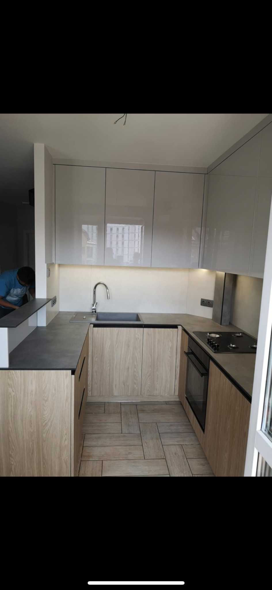 Nowoczesna kuchnia w kształcie litery U z drewnianymi frontami, szarymi blatami i połyskującymi szafkami górnymi. Widoczny fragment osoby pracującej w kuchni.