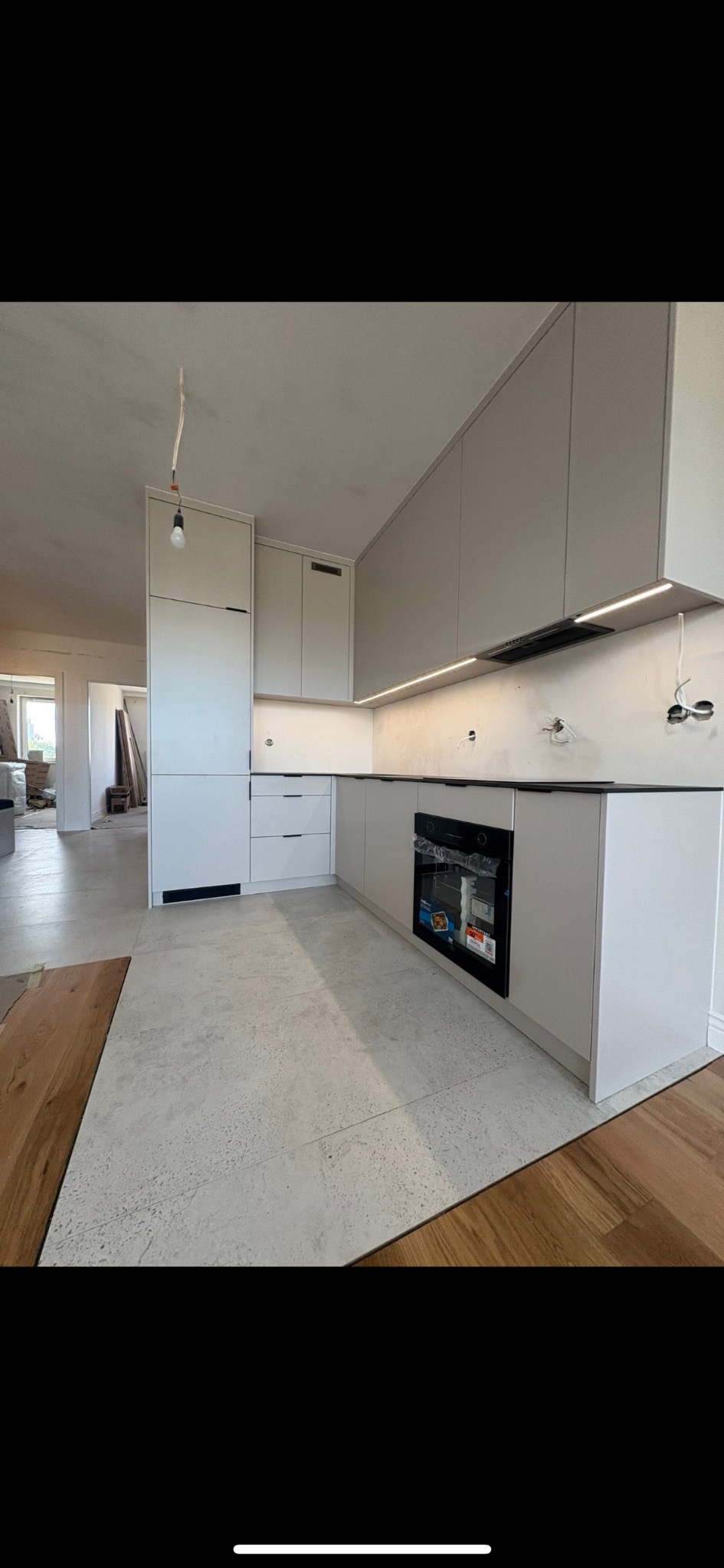 Jasna kuchnia na wymiar w trakcie montażu: białe szafki, czarny blat, piekarnik. Widoczne niedokończone instalacje elektryczne i surowa podłoga.