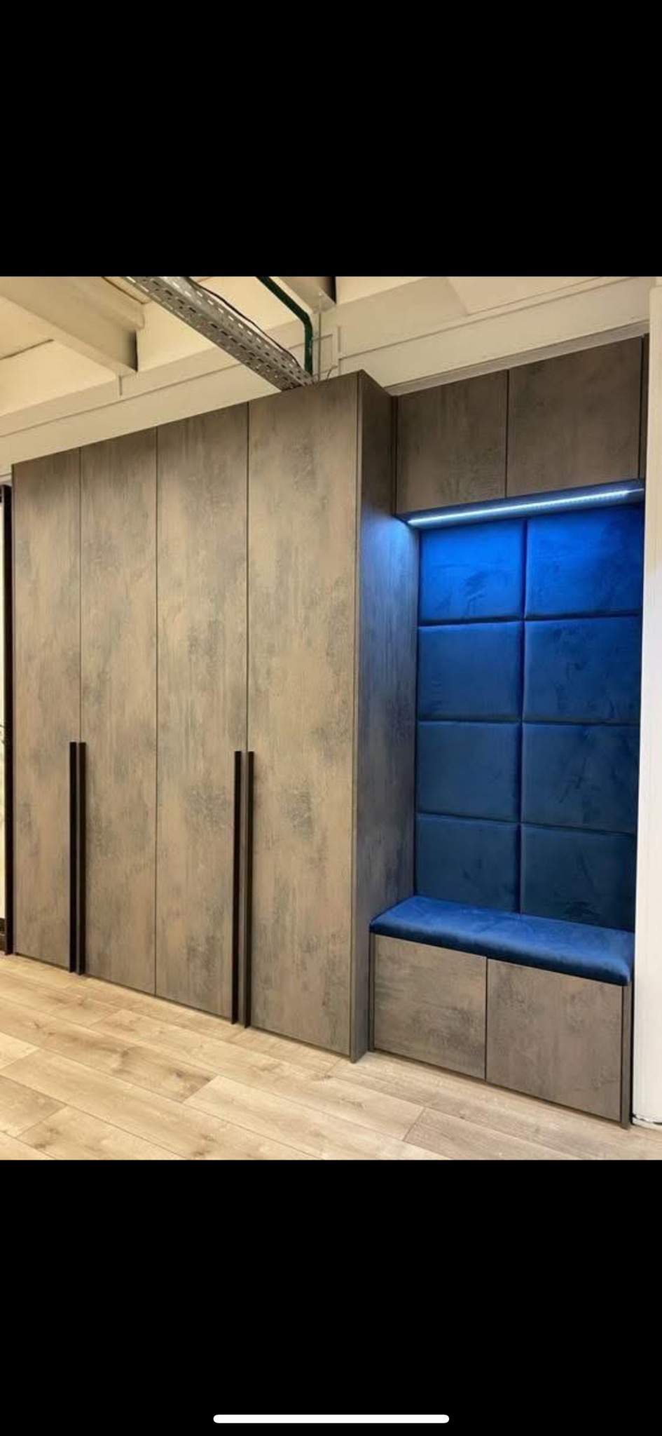 Szara szafa wnękowa z siedziskiem i niebieskim panelem tapicerowanym z oświetleniem LED, na jasnej podłodze z paneli. Minimalistyczny design.
