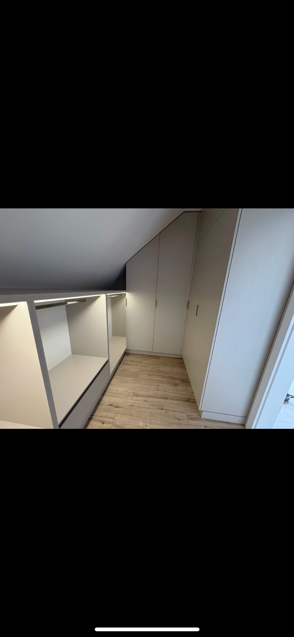 Jasna garderoba na poddaszu ze schowkami i szafami z frontami bez uchwytów. Oświetlenie LED podkreśla nowoczesny charakter wnętrza.