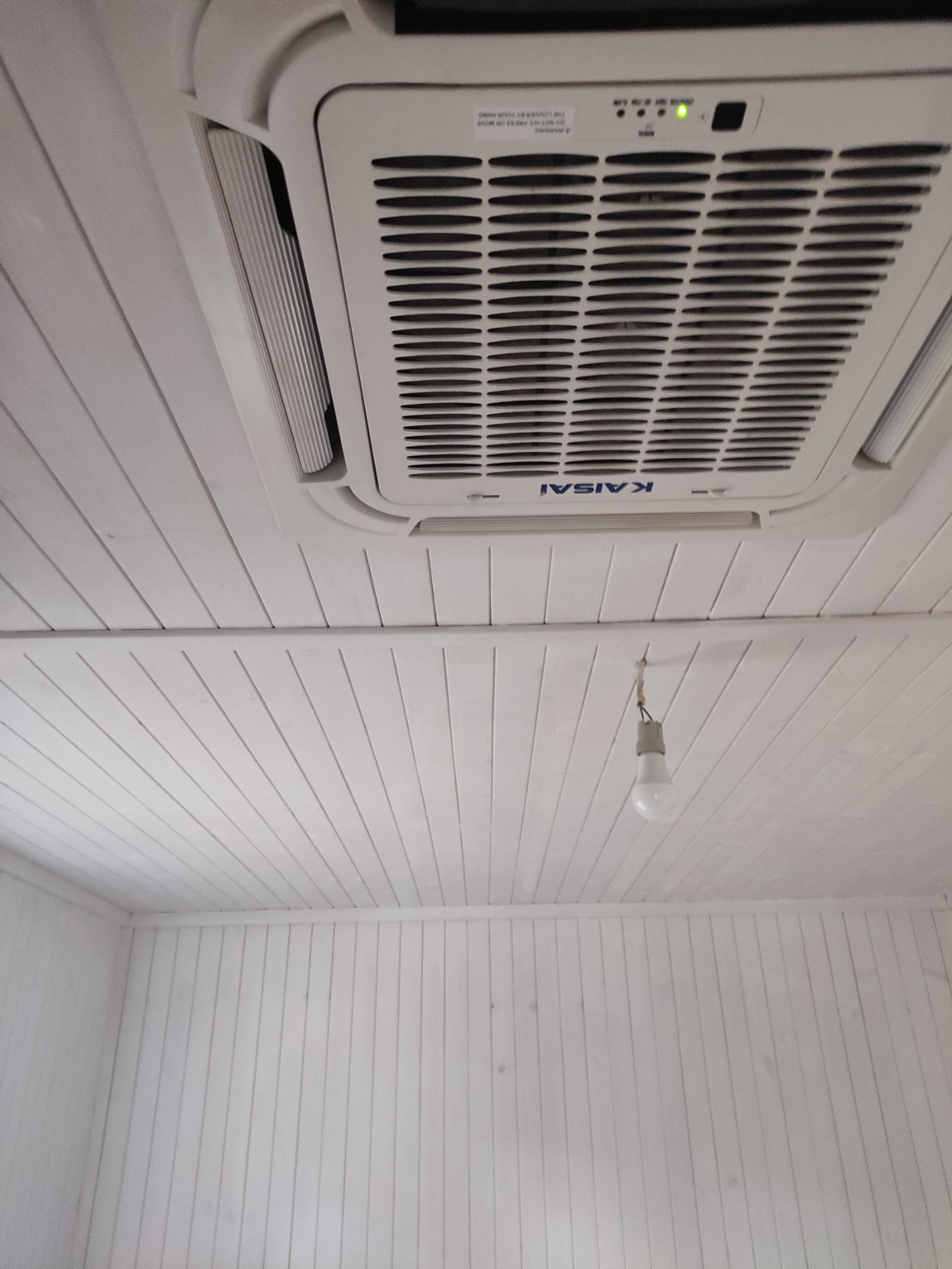 Białe, drewniane panele sufitowe z zamontowanym klimatyzatorem Kaisai i wiszącą żarówką. Widok z dołu, oświetlenie dzienne.