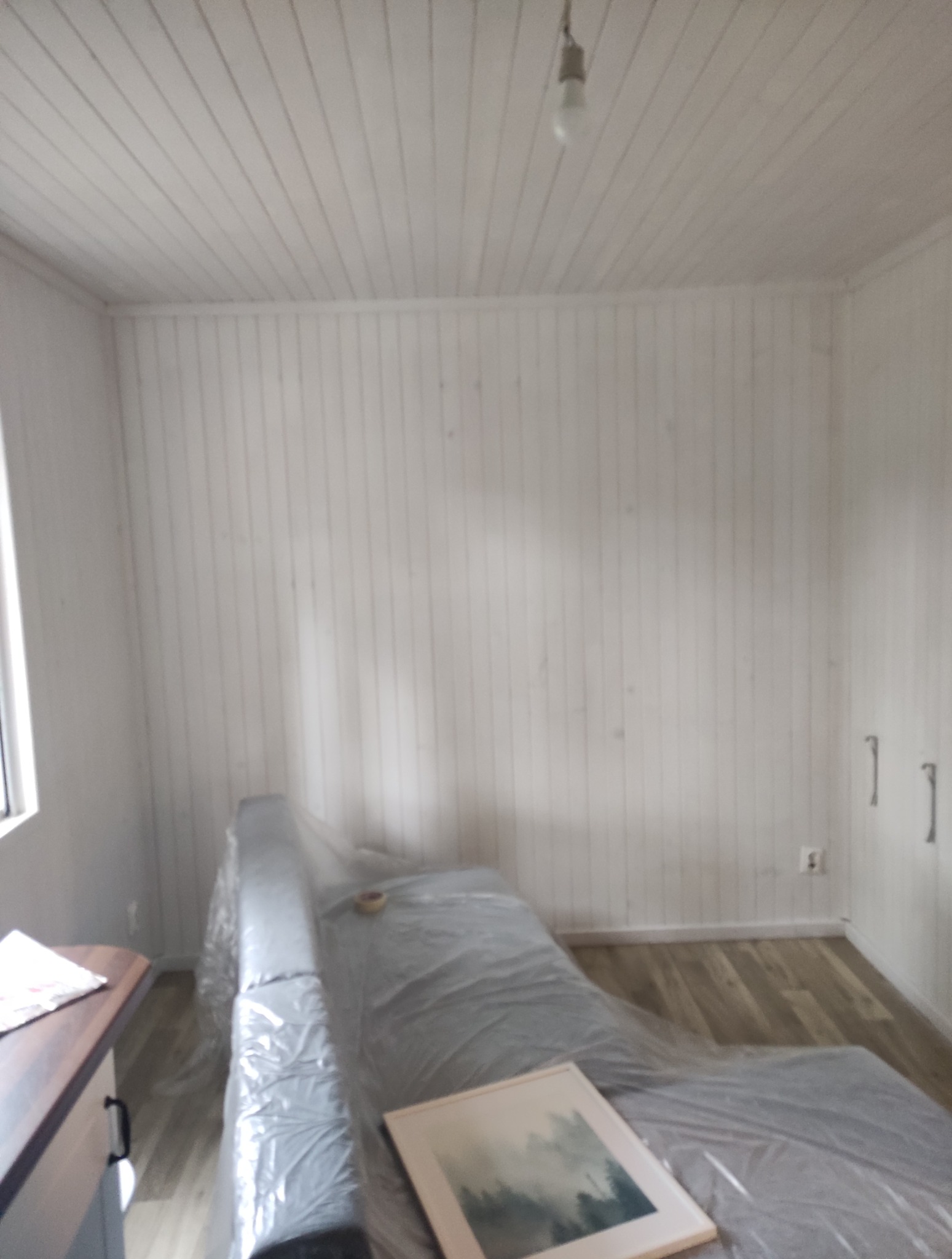 Białe panele ścienne i sufitowe w pokoju, zabezpieczone meble folią malarską, widoczna podłoga z paneli. Przygotowanie do odświeżenia wnętrza.