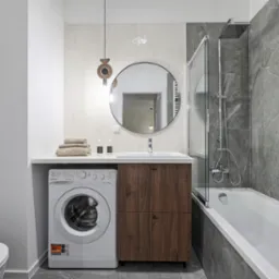 Nowoczesna łazienka z pralką obok szafki umywalkowej z drewnianymi frontami. Nad umywalką okrągłe lustro i designerska lampa. Szare płytki imitujące marmur.