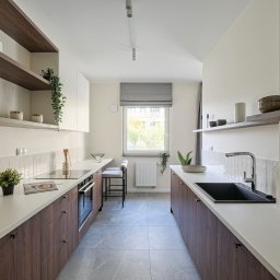 PauLine Concept Paulina Oszczyk - Nowoczesna kuchnia w odcieniach bieli i brązu. Minimalistyczny design z drewnianymi szafkami, białym blatem i szarymi płytkami. Okno z roletą rzymską w tle.