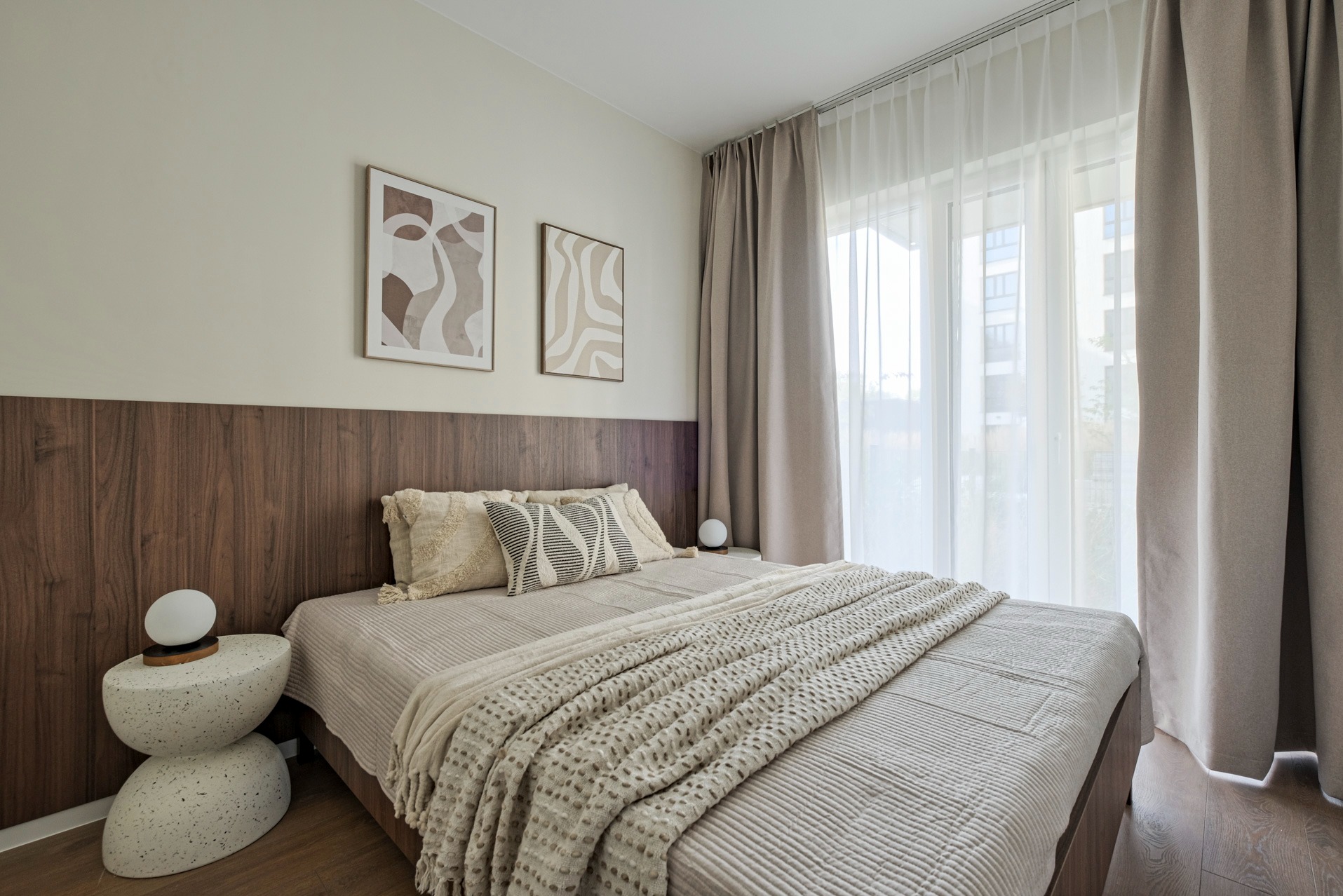 Jasna sypialnia z drewnianym panelem za łóżkiem, beżowymi zasłonami i abstrakcyjnymi obrazami. Nocna lampka na stoliku w kształcie kamieni.