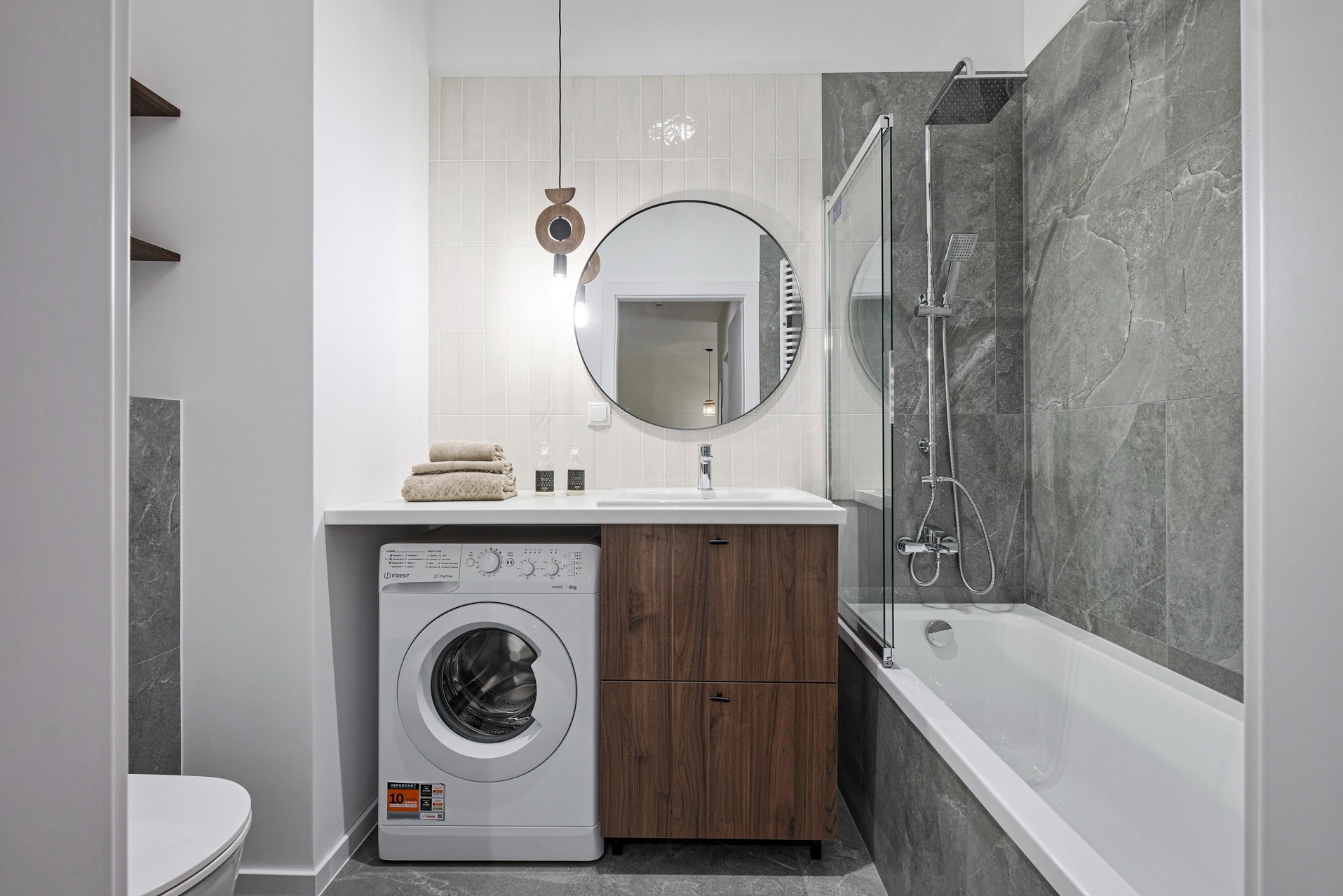 Nowoczesna łazienka z pralką obok szafki umywalkowej z drewnianymi frontami. Nad umywalką okrągłe lustro i designerska lampa. Szare płytki imitujące marmur.