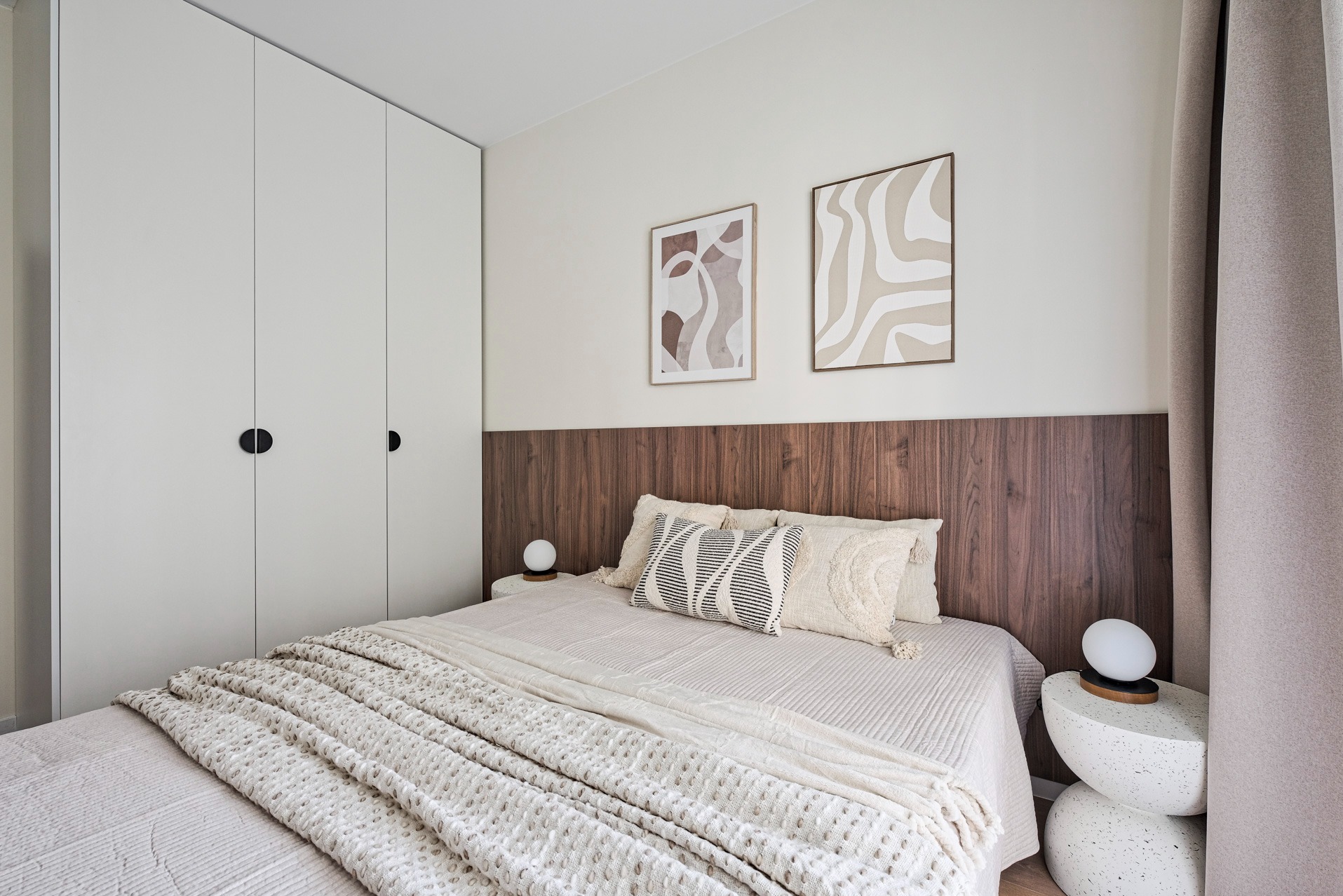 Minimalistyczna sypialnia z drewnianym zagłówkiem, beżową pościelą i białymi szafami. Dwie abstrakcyjne grafiki wiszą nad łóżkiem.