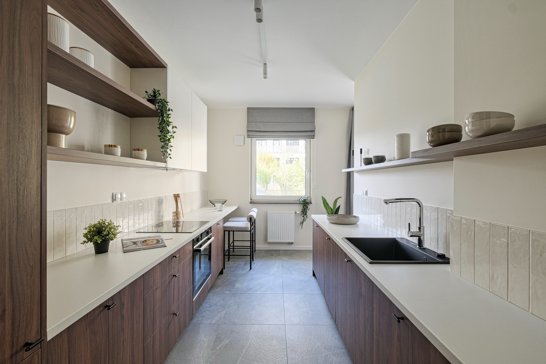 Nowoczesna kuchnia w odcieniach bieli i brązu. Minimalistyczny design z drewnianymi szafkami, białym blatem i szarymi płytkami. Okno z roletą rzymską w tle.