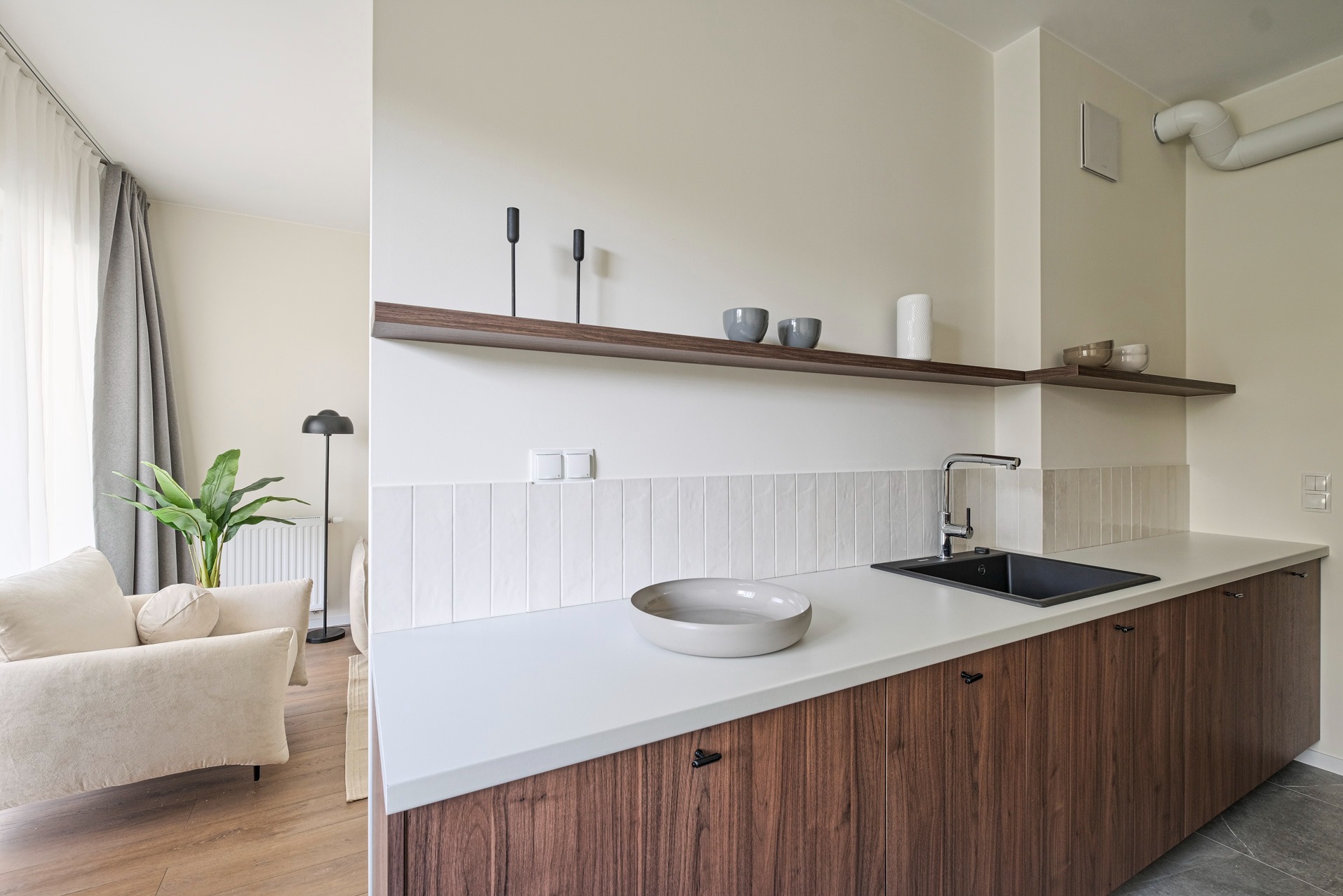 Minimalistyczna kuchnia z drewnianymi szafkami, białym blatem i płytkami. Na półkach dekoracje, w tle salon z beżową sofą i rośliną. Nowoczesne wykończenie wnętrza.