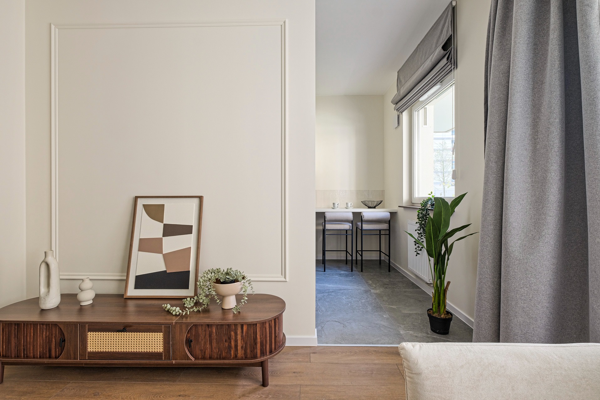 Stylowe wnętrze: drewniana komoda z dekoracjami, abstrakcyjny obraz w ramie, rośliny doniczkowe i szare zasłony w tle, tworzące spójną aranżację.