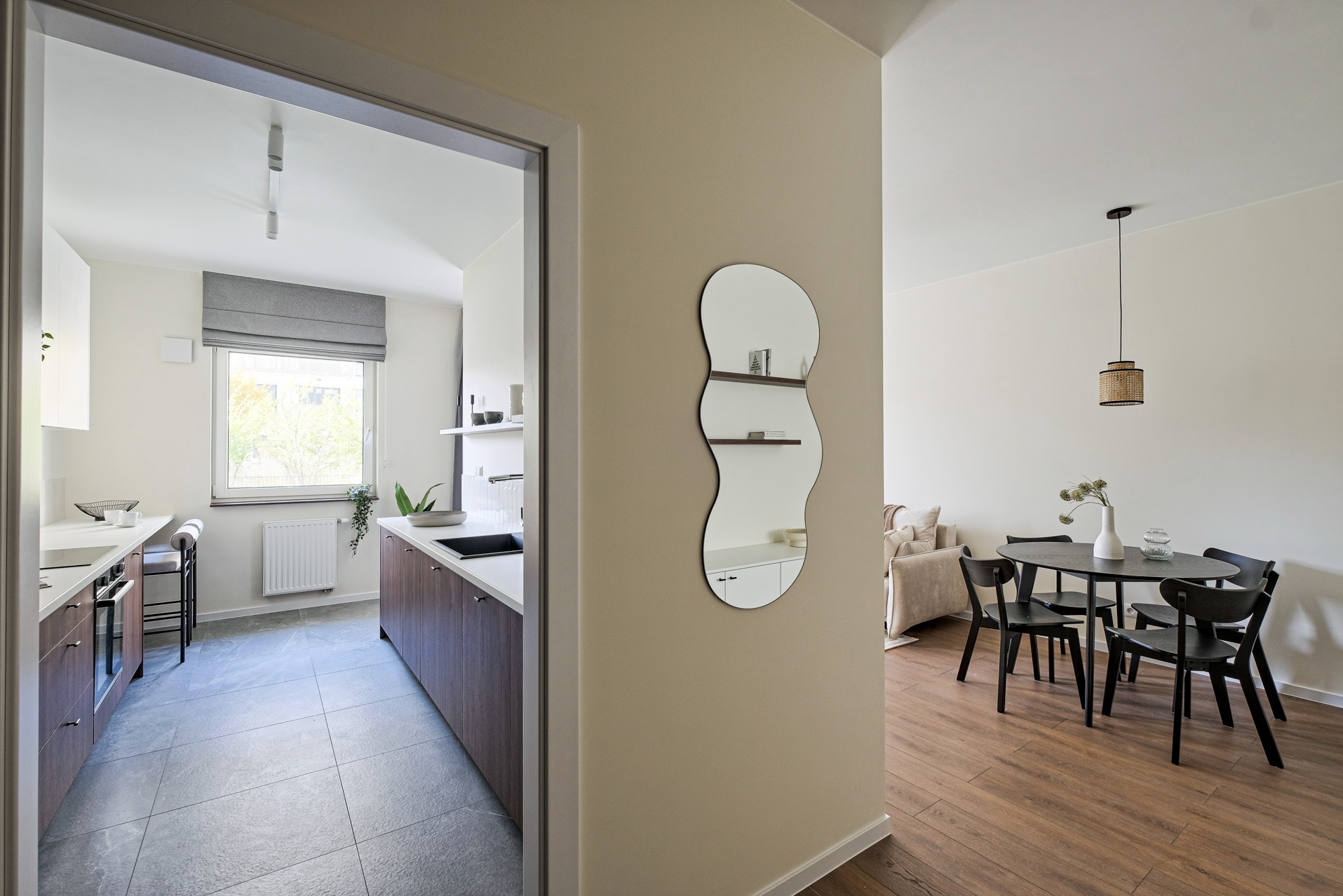 Nowoczesne wnętrze: kuchnia z ciemnymi szafkami, jadalnia z czarnym stołem i krzesłami, oraz designerskie lustro na ścianie. Widok z perspektywy drzwi.