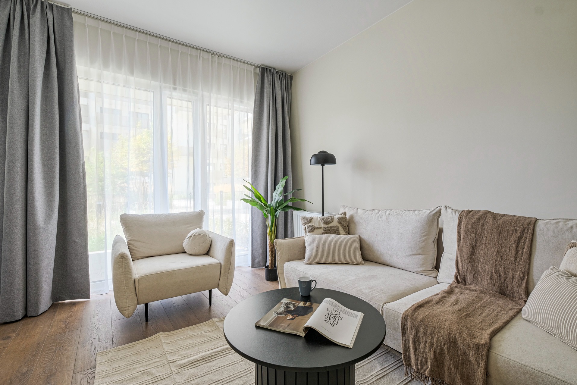 Jasne wnętrze salonu z beżową sofą, fotelem i okrągłym stolikiem kawowym. Duże okno z zasłonami w odcieniach szarości. Na stoliku leży otwarta książka.
