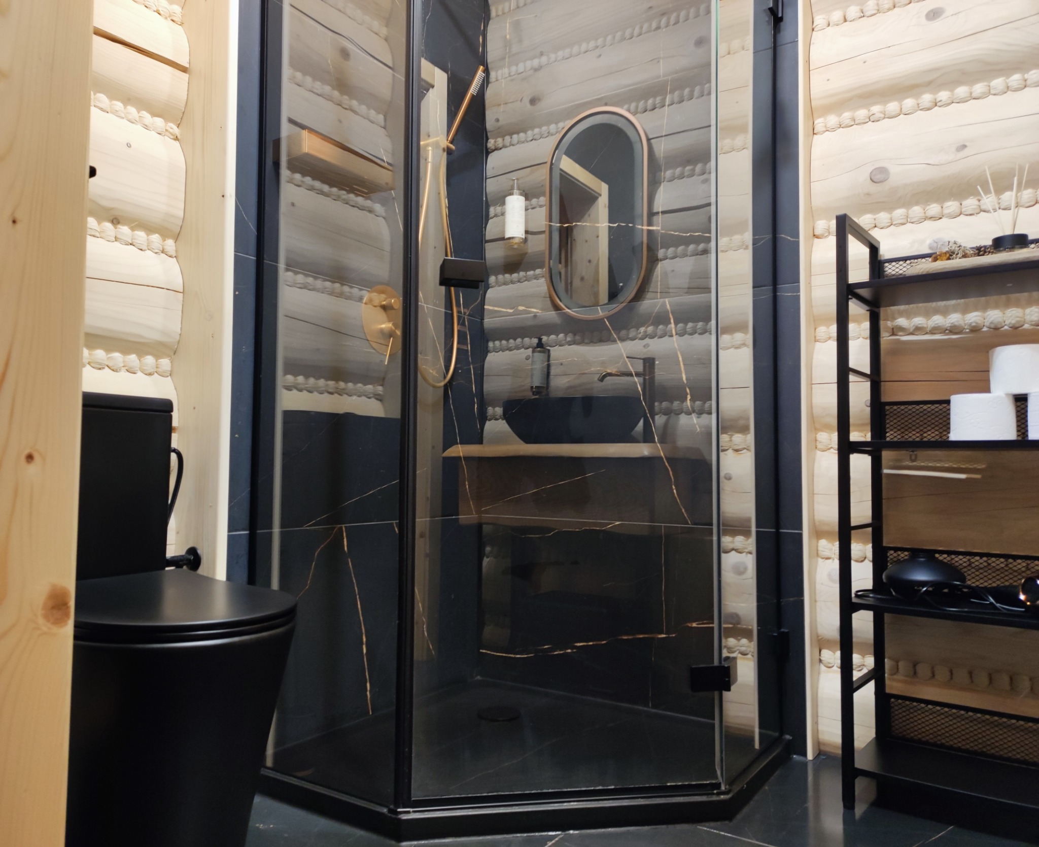 Nowoczesna łazienka w Istebnej z kabiną prysznicową typu walk-in, czarną armaturą i drewnianymi elementami wykończeniowymi. Widoczna toaleta i półka z akcesoriami.