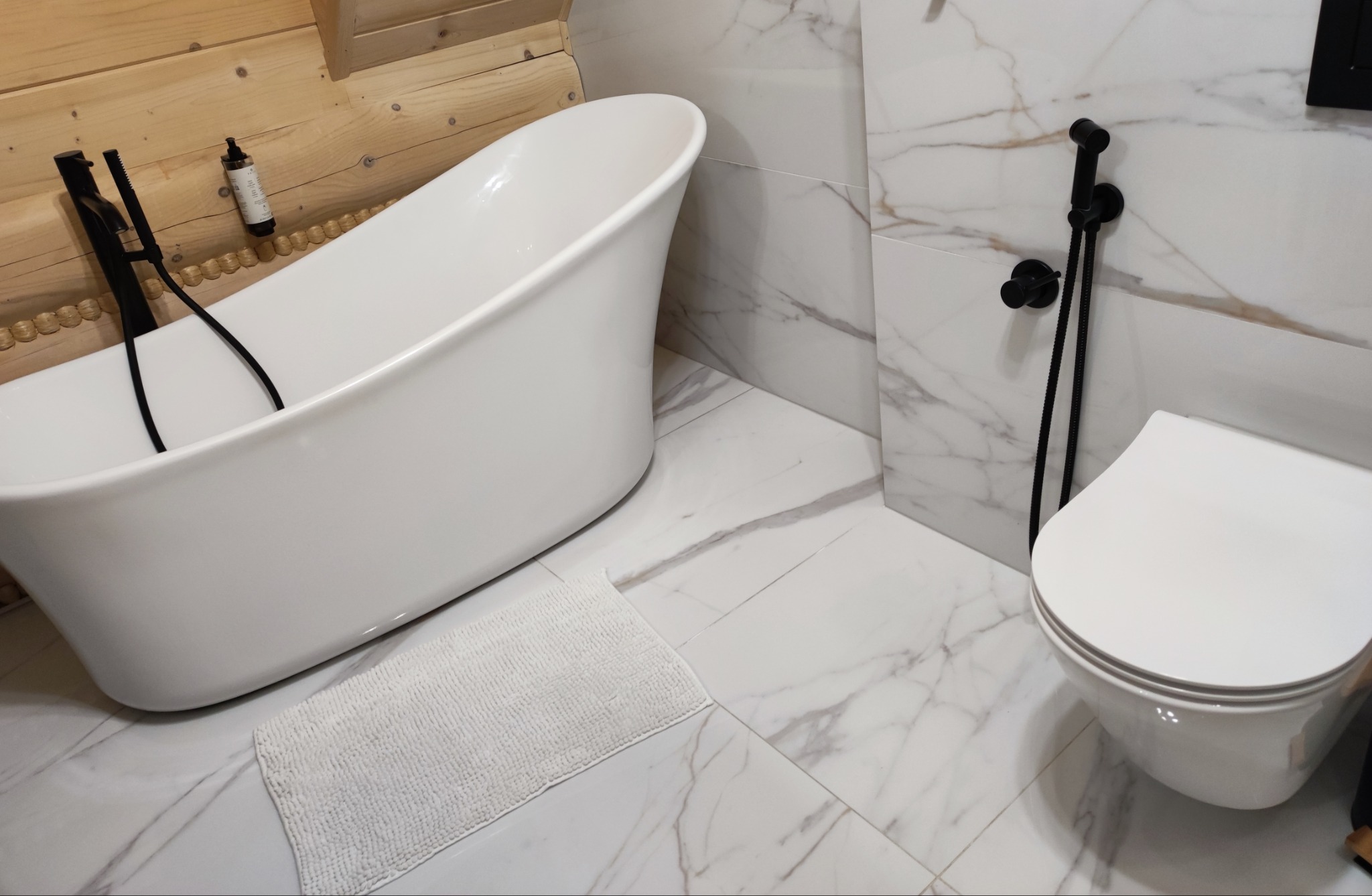 Nowoczesna łazienka z wolnostojącą wanną, białą armaturą i marmurowymi płytkami. Czarna bateria wannowa i bidetta kontrastują z jasnym wnętrzem. Drewniane elementy dodają ciepła.