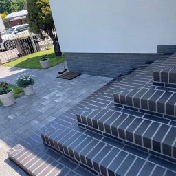 Mateusz Leśnikowski - Schody wejściowe wyłożone ciemną, ceglastą kostką brukową, prowadzące do białego budynku. Widoczne donice z kwiatami i fragment posesji z samochodem.