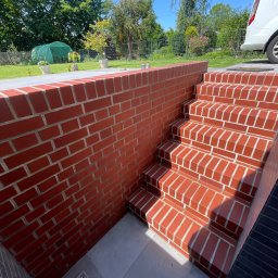 Mateusz Leśnikowski - Ceglane schody zewnętrzne z jasnymi fugami, prowadzące w dół, obok muru z czerwonej cegły. Widoczny fragment trawnika i roślinność w tle, w słoneczny dzień.