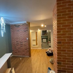 Mateusz Leśnikowski - Ceglana ściana działowa w odcieniach czerwieni, drewniana podłoga, widok na dalsze pomieszczenia. Dekoracyjne oświetlenie sufitowe. Częściowo widoczne przedmioty w pomieszczeniu.